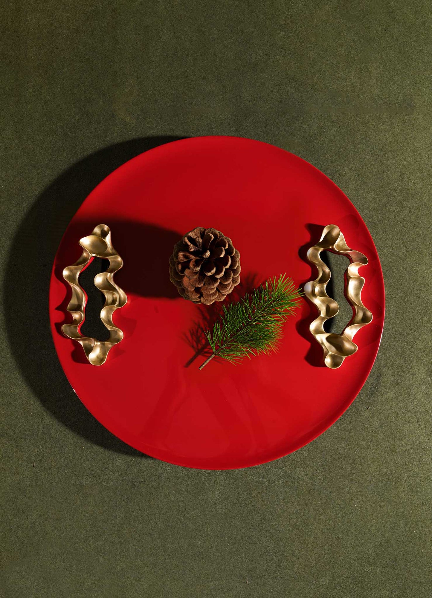 A Saffron Red Tray Small by VIYA rests on a dark surface, displaying a pine cone, a small pine branch, and two bronzed objects arranged symmetrically.