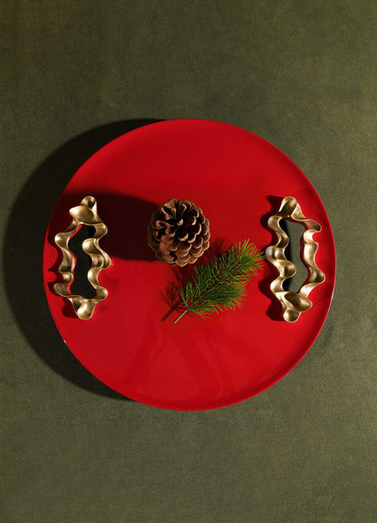 A Saffron Red Tray Small by VIYA rests on a dark surface, displaying a pine cone, a small pine branch, and two bronzed objects arranged symmetrically.
