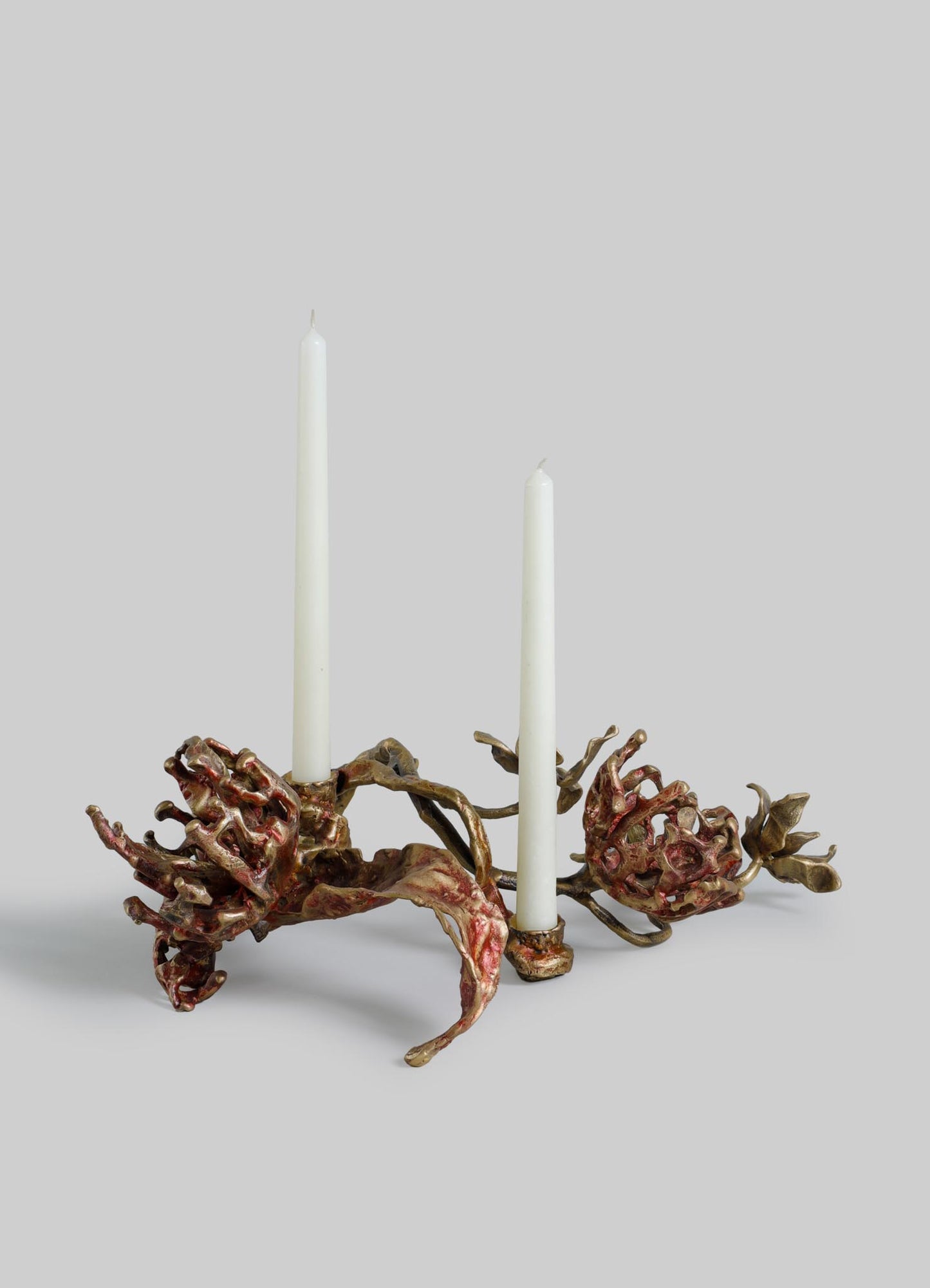 A sculptural candle holder with an organic, floral-inspired design in a metallic bronze and red finish, featuring two white taper candles.