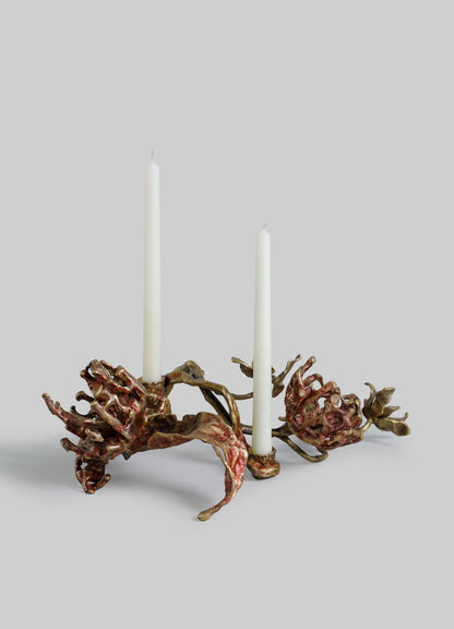 A sculptural candle holder with an organic, floral-inspired design in a metallic bronze and red finish, featuring two white taper candles.