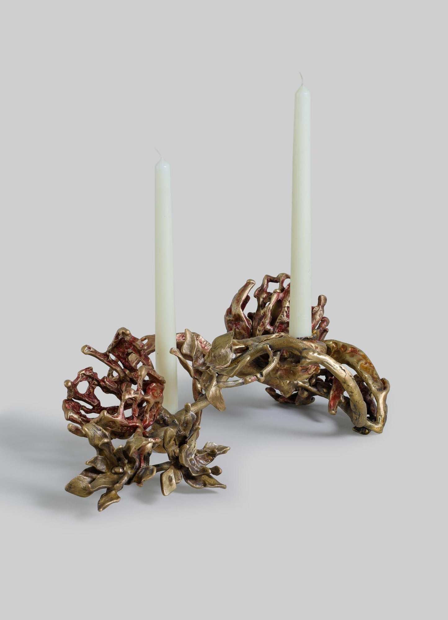 A decorative candle holder crafted from metal with an organic, floral-inspired design in bronze and reddish tones, holding two tall white candles. The piece has a handcrafted style with intricate, flowing motifs resembling leaves and flowers.