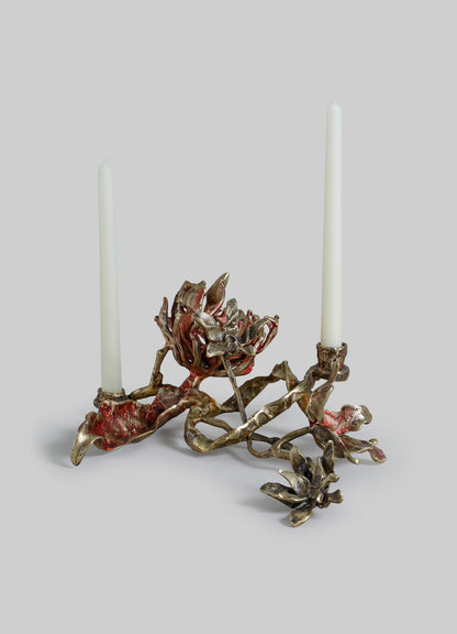 A decorative candle holder with a sculptural, botanical motif in a metallic finish, featuring two white taper candles with a rustic, artistic design.