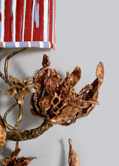 A close-up of the VIYA Spring In Kashmir Wall Sconce A, featuring a sculpted metallic floral design with patina finish and a red, white, and blue striped shade against a plain light gray wall for a Kashmir-inspired home decor look.