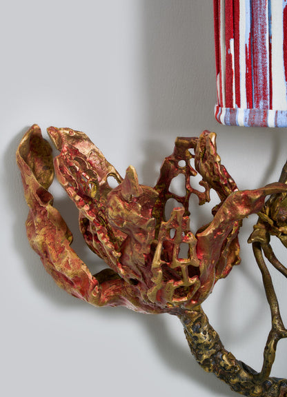 Close-up of VIYA's "Spring In Kashmir Wall Sconce B," a sculpted brass and bronze metallic flower in red and gold tones on a branch, set against red, white, and blue striped fabric. Ideal for Kashmir-inspired home decor.