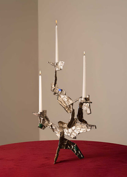 The VIYA Tutti Frutti Nickel Candelabra, featuring an abstract angular design with embedded semi-precious stones and three white candles, stands on a red velvet surface against a plain beige background.