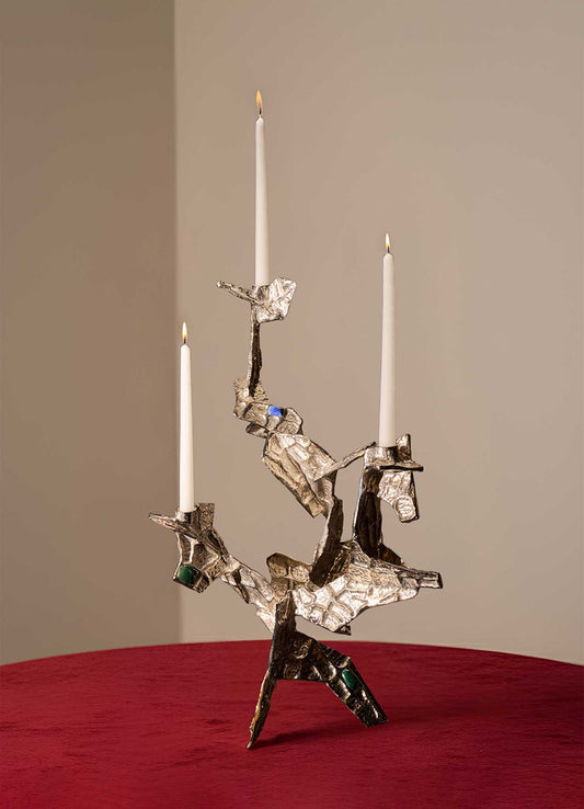 The VIYA Tutti Frutti Nickel Candelabra, featuring an abstract angular design with embedded semi-precious stones and three white candles, stands on a red velvet surface against a plain beige background.