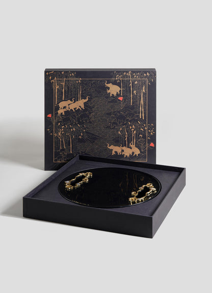 The VIYA Saffron Ecru Tray Large is a round, black tray with gold animal designs, presented in an open box. Its lid displays matching gold animal and forest motifs on a dark background with subtle red accents.
