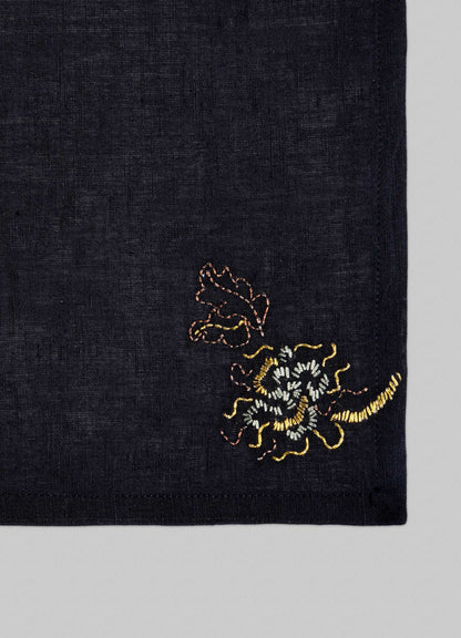The Viya Tree Full of Parrots Hand Embroidered Black & Gold Cocktail Napkins (Set of 4) feature dark linen with small yellow and brown floral embroidery in one corner, set against a plain light grey background.