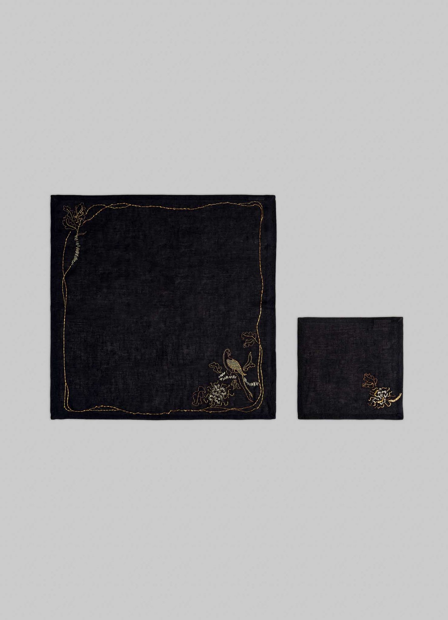 The Viya Tree Full of Parrots Hand Embroidered Black & Gold Napkin features two black napkins with gold floral embroidery in one corner—one folded, one unfolded—displayed on a light gray background, symbolizing blessings and good fortune.
