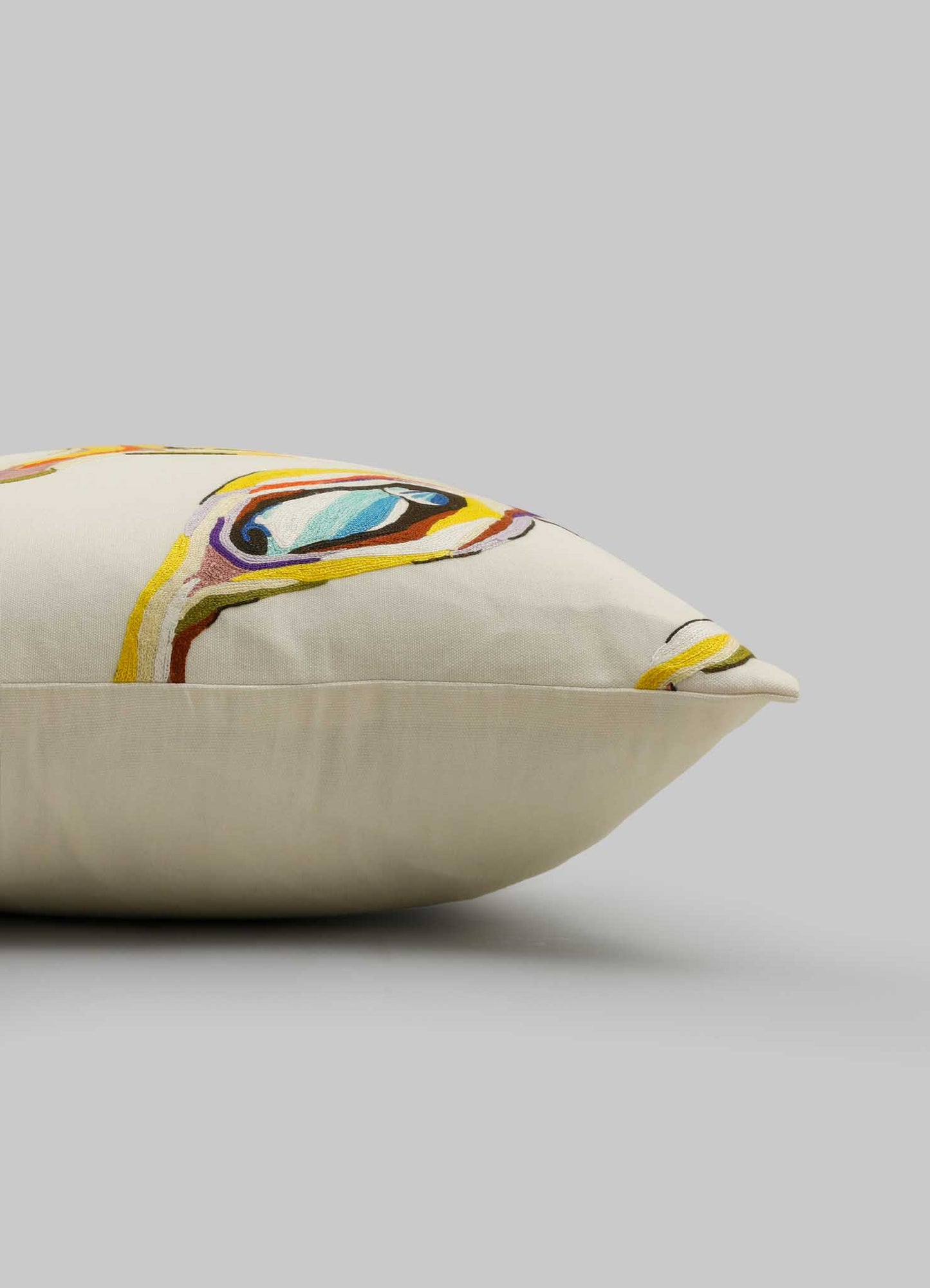 The Viya Tutti Frutti Hand Embroidered Multicolour Cushion Cover (18"x18") features a cream base with vibrant blue, yellow, and purple abstract stitching that resembles an animal face, displayed against a plain gray background.