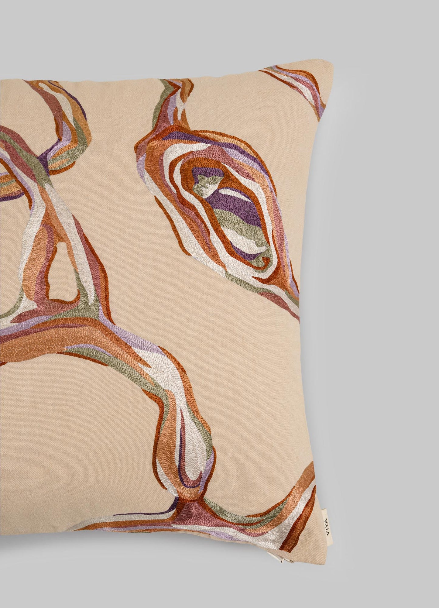 The Viya Tutti Frutti Salmon Pink Cushion Cover (18"x18") features abstract wavy embroidery in brown, orange, purple, and white on a light gray background with a small fabric tag in the bottom right corner.