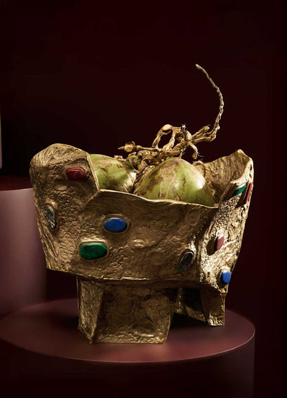 A decorative bowl with a textured gold finish and colorful gemstone accents, filled with green fruits and dried branches, placed on a round pink surface against a dark background.