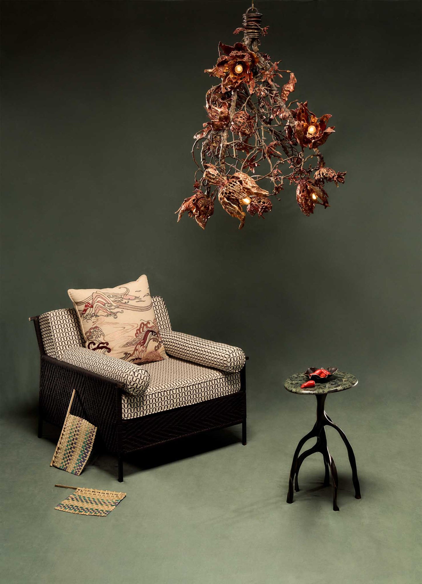 A cozy armchair with patterned cushions sits beside a small round table topped with red berries, beneath the VIYA Spring In Kashmir Chandelier, inspired by Kashmir decor and decorative patinas, set against a plain green background.