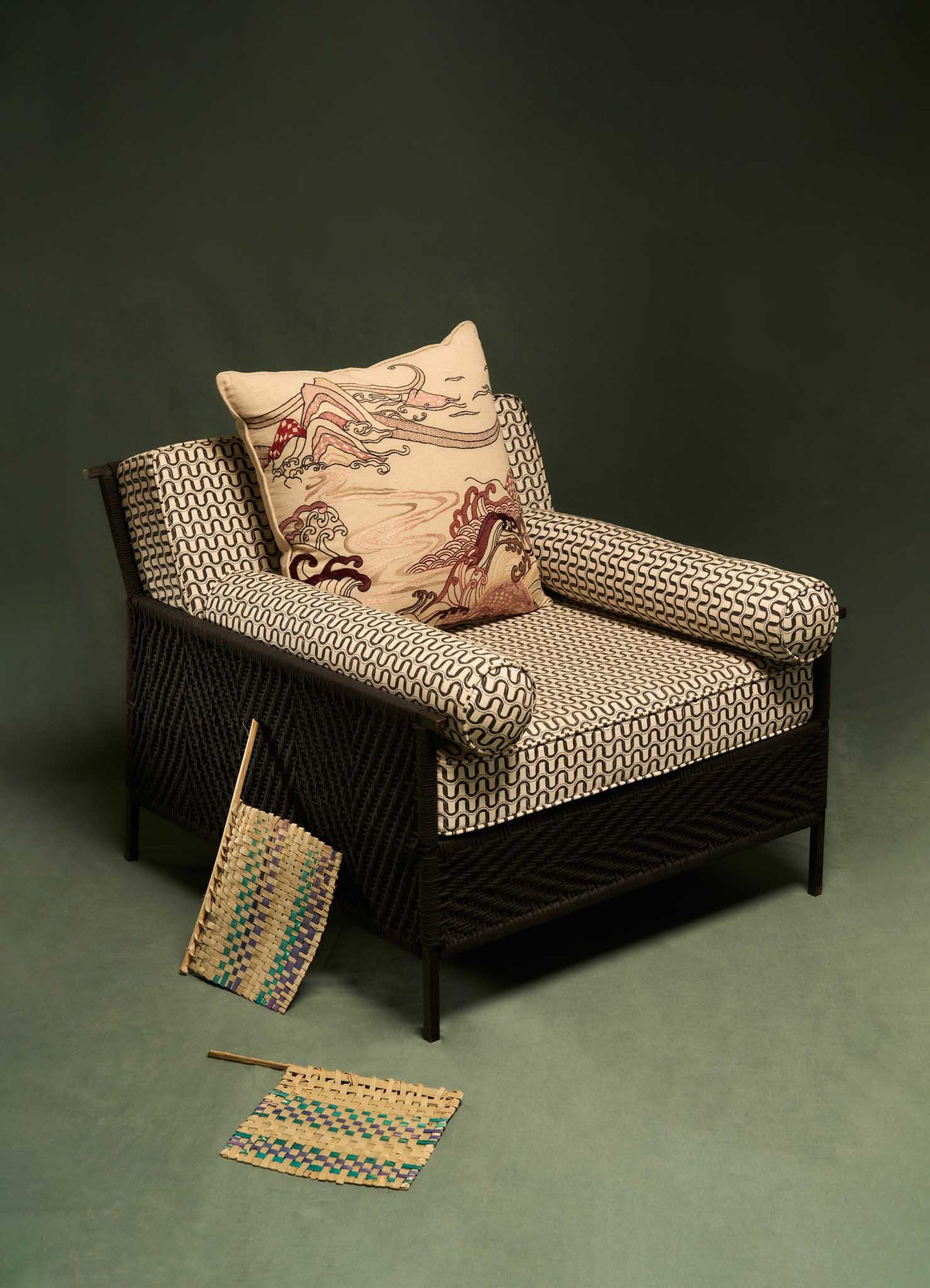Wicker armchair with black woven texture and patterned fabric cushions featuring geometric black and white designs; includes a decorative pillow with a detailed illustration of a landscape and woven mats with multicolored accents, on a green floor.